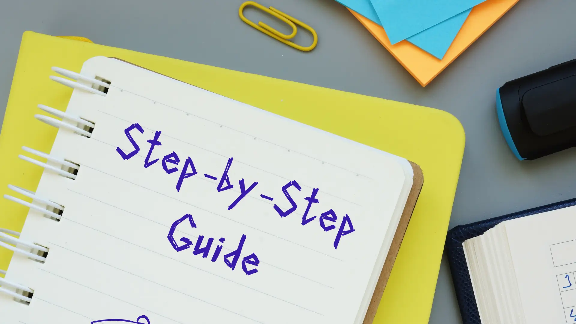 Close‑up of spiral notebook with handwritten title “Step‑by‑Step Guide” in purple text, placed on yellow folder with surrounding office supplies