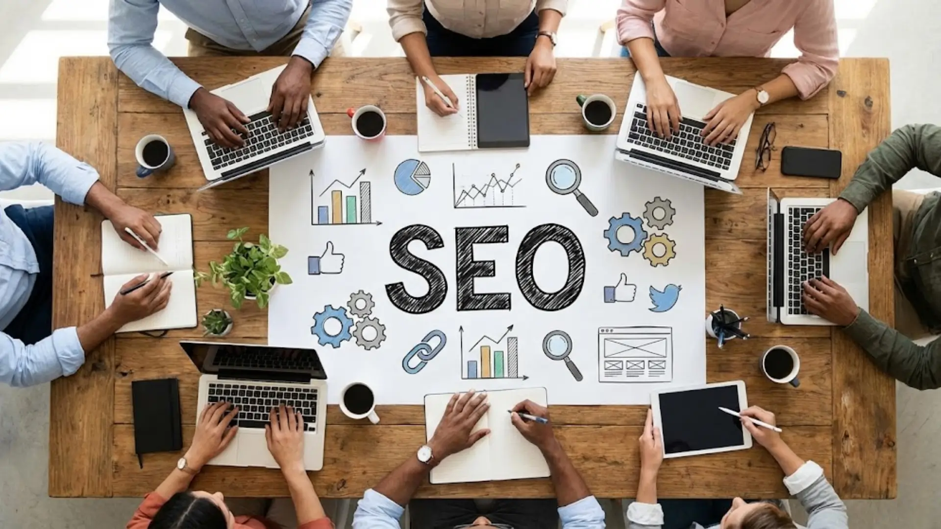 Top-down view of a team working around a wooden table with laptops, tablets, notebooks, and coffee cups. A large sheet in the center shows the word ‘SEO’ surrounded by icons for charts, gears, magnifying glass, social media, and links, representing collaborative search engine optimization work.