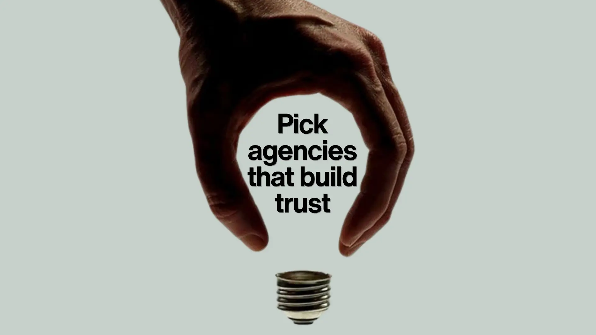 Hand positioned as if holding the top of a light bulb, with text inside reading “Pick agencies that build trust.” Symbolic composition linking trust with ideas and clarity.