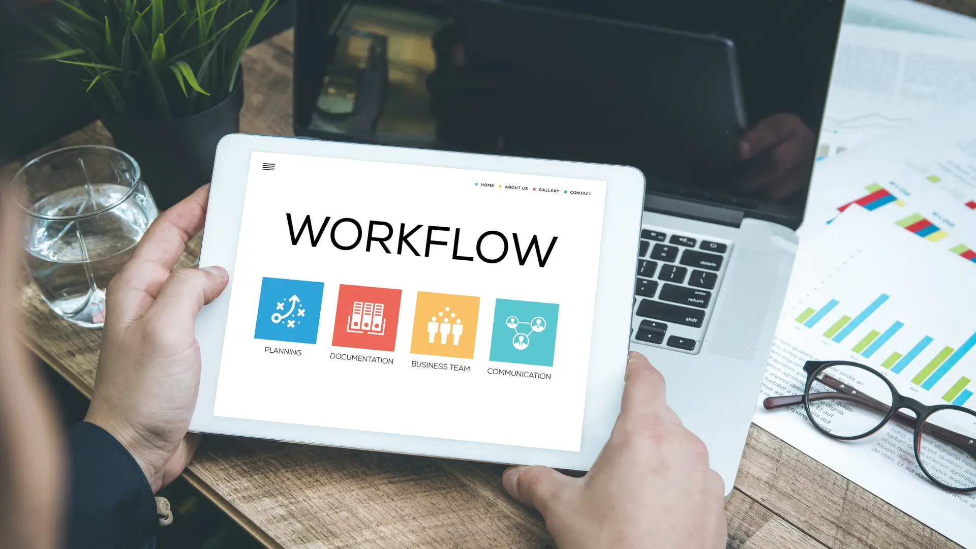 Person holding tablet displaying workflow interface with icons for planning, documentation, business team, and communication, over desk with laptop, eyeglasses, charts, and water glass.
